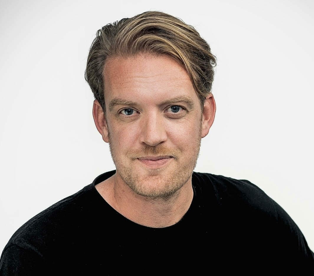 A man with light brown hair and stubble, wearing a black t-shirt, looks directly at the camera against a plain white background, embodying the innovative spirit of Spryker's ShoppingTomorrow initiative.