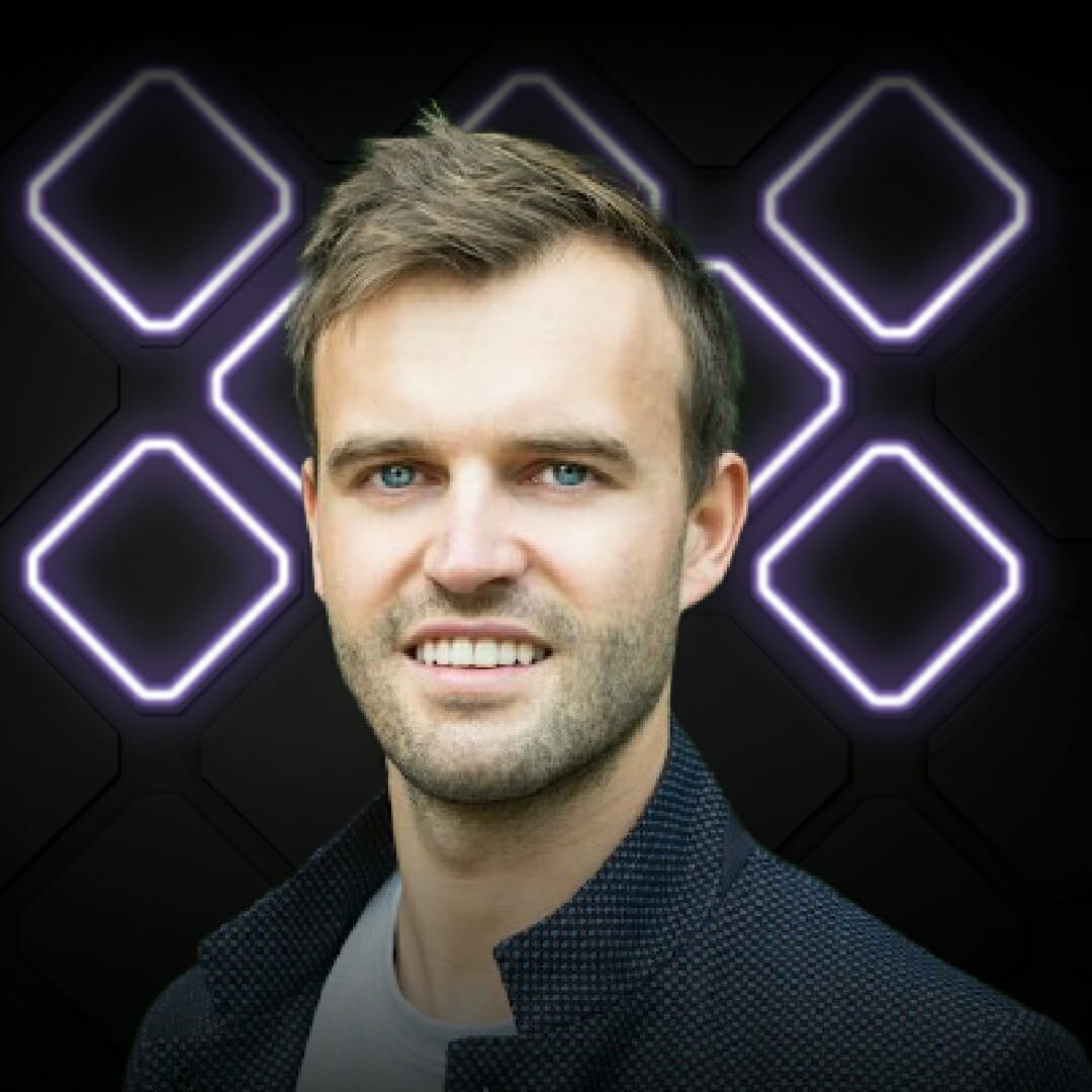 A man with short hair and light stubble wearing a dark jacket and white shirt is standing in front of a background with glowing geometric shapes, capturing the vibrant energy of Spryker EXCITE 2022.