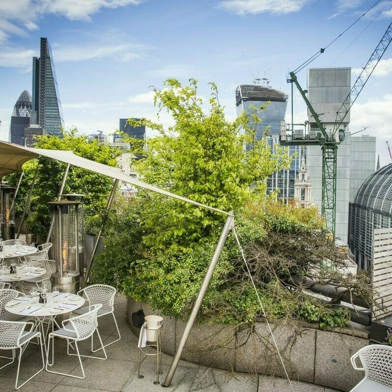 Rooftop outdoor dining area with white tables and chairs, shaded by a tent, overlooking city skyscrapers, trees, and a construction crane in the background—ideal for discussing innovations like Spryker at Shoptalk Europe.