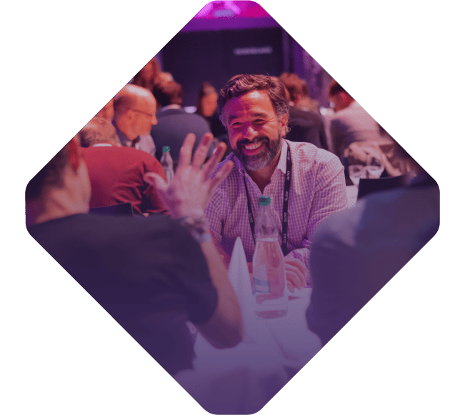 A group of people, including a bearded man smiling, engage in conversation at a table during the Spryker EXCITE 2022 event.