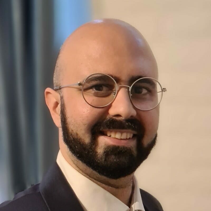 A bald man with glasses, a beard, and a mustache is smiling while wearing a dark suit and white shirt, possibly attending a customer advisory board meeting.