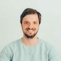 A man with short brown hair and a beard wearing a light blue sweater smiles at the camera against a plain white background, embodying the friendly spirit of Spryker careers.