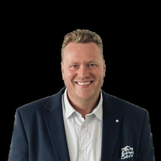 A man with short blonde hair, wearing a dark blazer and white shirt, is smiling against a plain black background—embodying the professional spirit of a customer advisory board member.