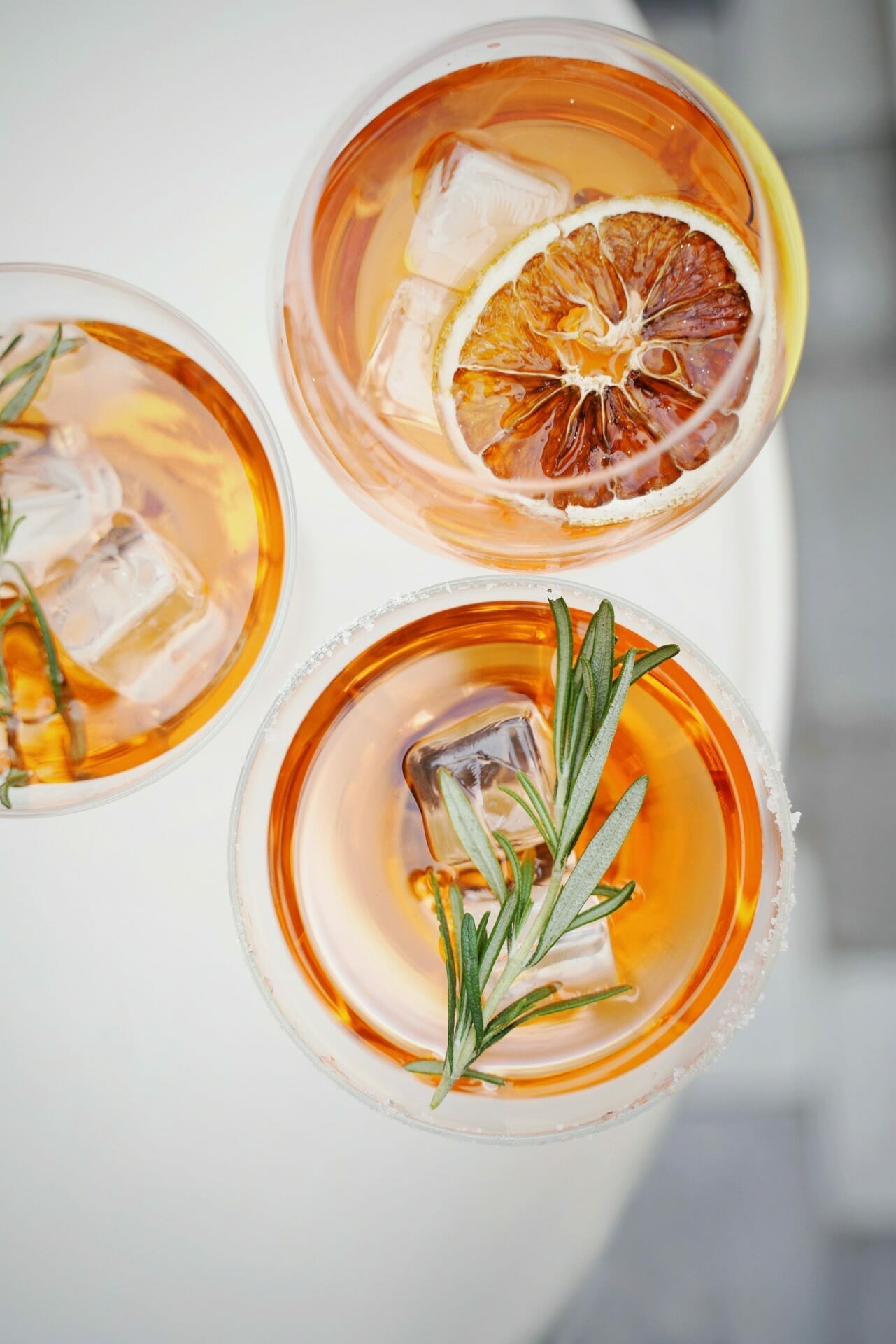 Three glasses of iced amber-colored drinks, garnished with slices of dried orange and sprigs of rosemary, viewed from above—a refreshing scene reminiscent of a toast at Spryker at Shoptalk Europe.