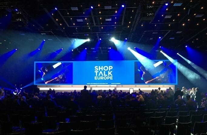 A large conference room with a stage displaying "ShopTalk Europe" on a large screen. Blue spotlights illuminate the stage while attendees sit and stand, engaging in conversations about Changing Gears 2022.