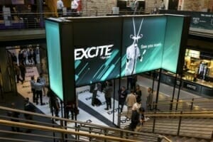 In a bustling Chicago shopping center, a large digital display boldly lights up with the word "EXCITE." Shoppers walk around, engaging with the dynamic B2B online promotions and exploring nearby stores.