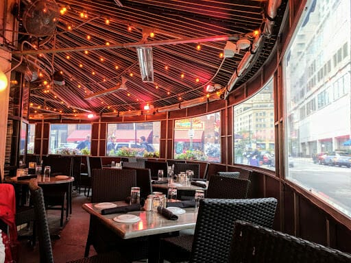 Outdoor restaurant patio with empty tables and chairs. The space is covered with a canopy and illuminated by string lights. Large windows offer a view of the bustling Chicago city street outside, providing an ideal B2B setting for networking or hosting online meetings in a vibrant setting.