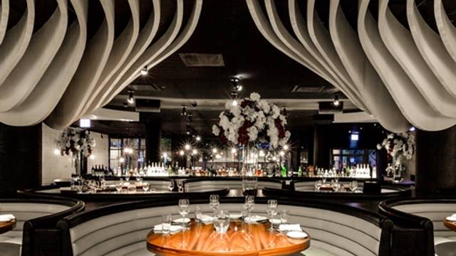 A modern restaurant interior with curved booth seating, a central round table set for four, and decorative white ceiling features sets the scene for B2B online meetings. A bar and illuminated shelves provide a chic backdrop in this Chicago dining hotspot.