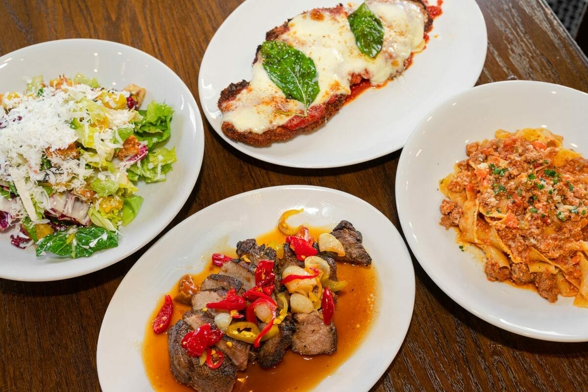 Four dishes on a wooden table: a salad with grated cheese, a breaded meat dish with melted cheese and basil, a pasta dish with meat sauce, and a plate of steak with peppers in sauce—a business lunch spread that could impress any B2B online meeting in Chicago.