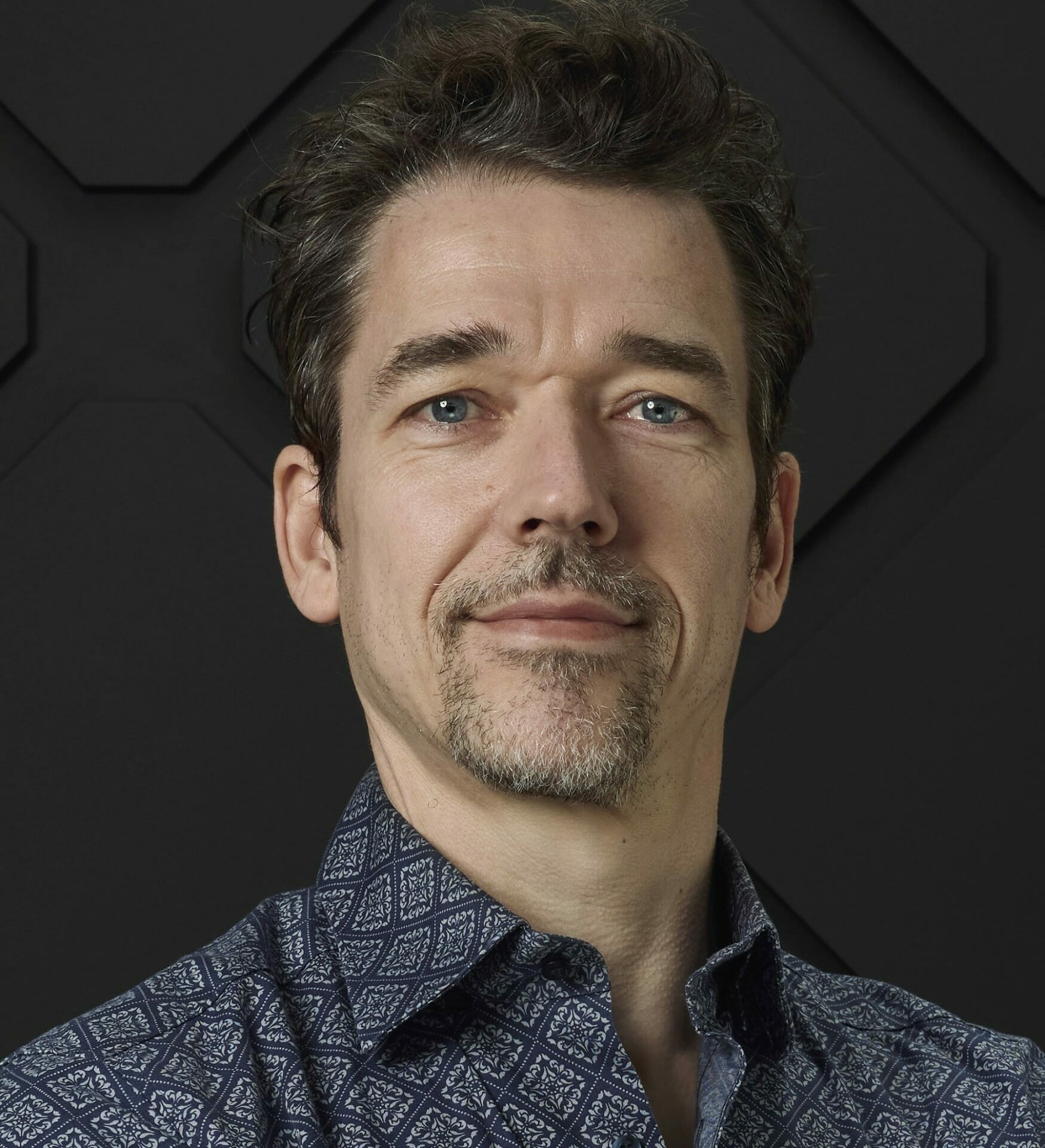 A man with short brown hair, a beard, and a mustache, wearing a patterned shirt, poses against a black geometric background at Shoptalk Las Vegas 2023.