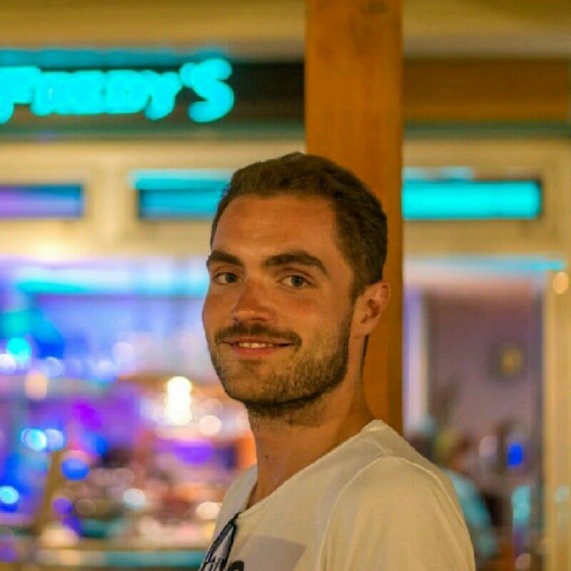 A man with short hair and a beard smiles at the camera. He is wearing a white shirt, and the background includes a brightly lit bar or restaurant sign, suggesting he might be out with his customer advisory board.