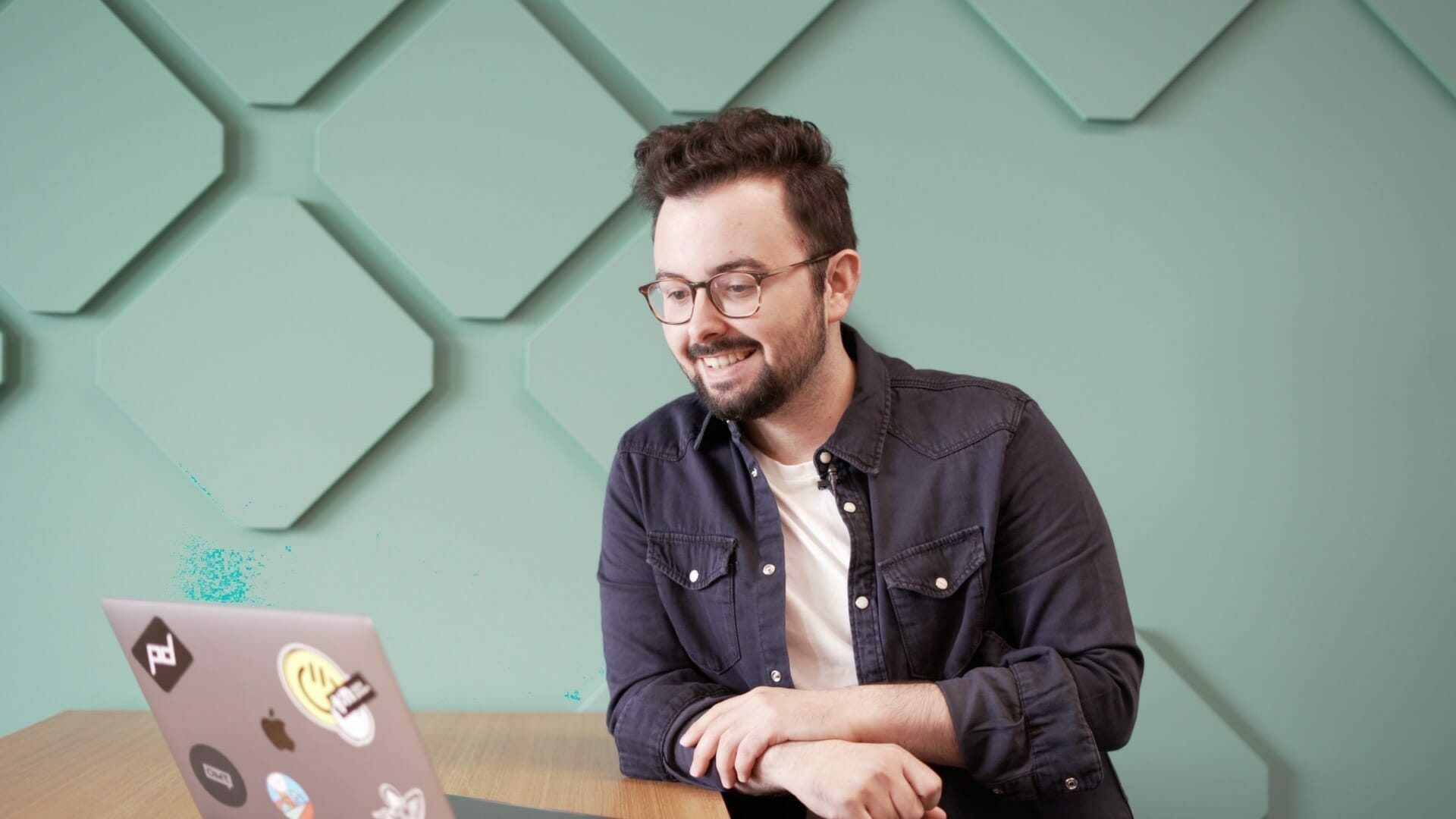 A man with glasses and a beard, wearing a dark shirt over a white t-shirt, is smiling and looking at a laptop on a wooden table against a geometric patterned green wall, possibly preparing for his Customer Advisory Board meeting.