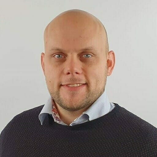 A bald man with a beard and mustache, wearing a dark sweater over a light blue collared shirt, smiles confidently in front of a plain white background—embodying the professional spirit of the B2B marketplace for Nordics.