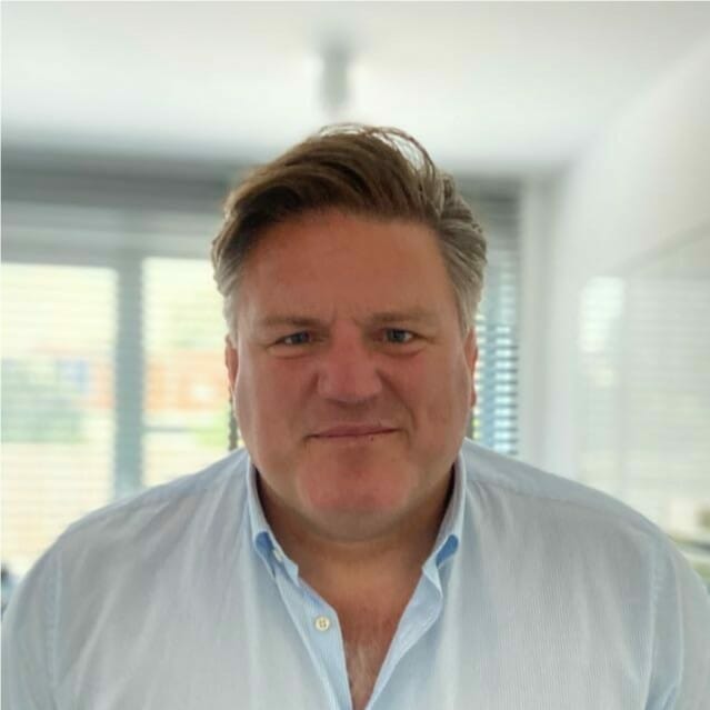 A man with short, light brown hair is standing indoors, wearing a light blue collared shirt. The background shows window blinds and a softly lit room, creating an ambiance perfect for Emerce e-commerce discussions.