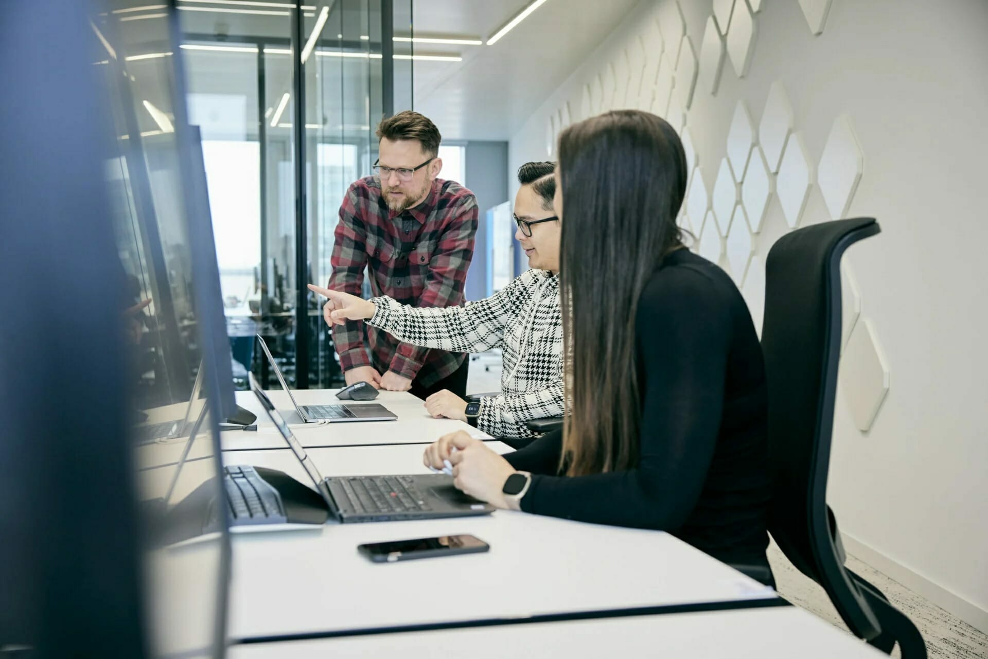 Three individuals in an office setting work together at a desk with laptops and monitors. One person points at a screen, demonstrating updates to the career page, while the others focus on the task.