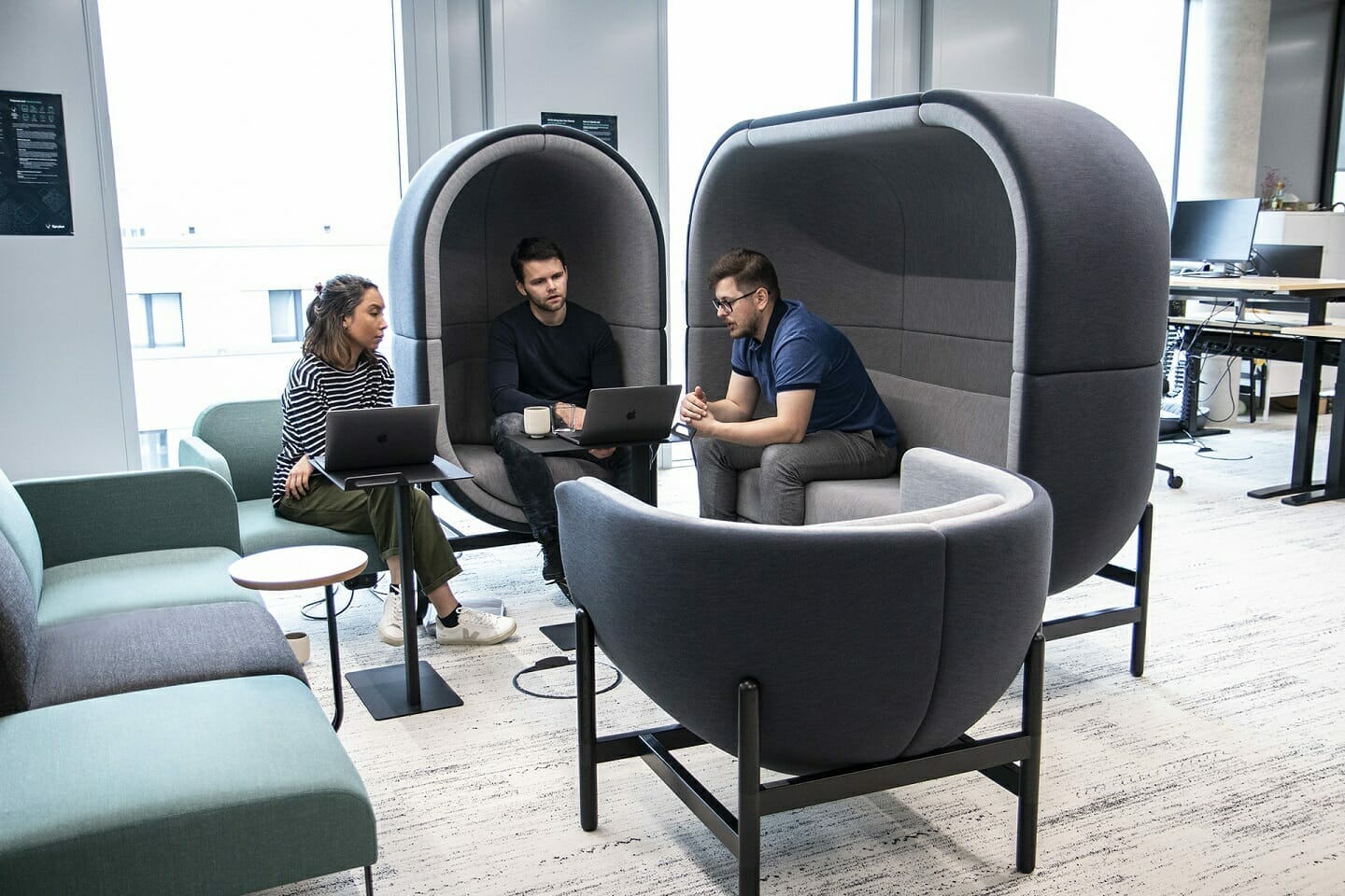 Three people are seated in modern, pod-like chairs, working on laptops and engaged in a discussion about Spryker careers in a bright, open office space.