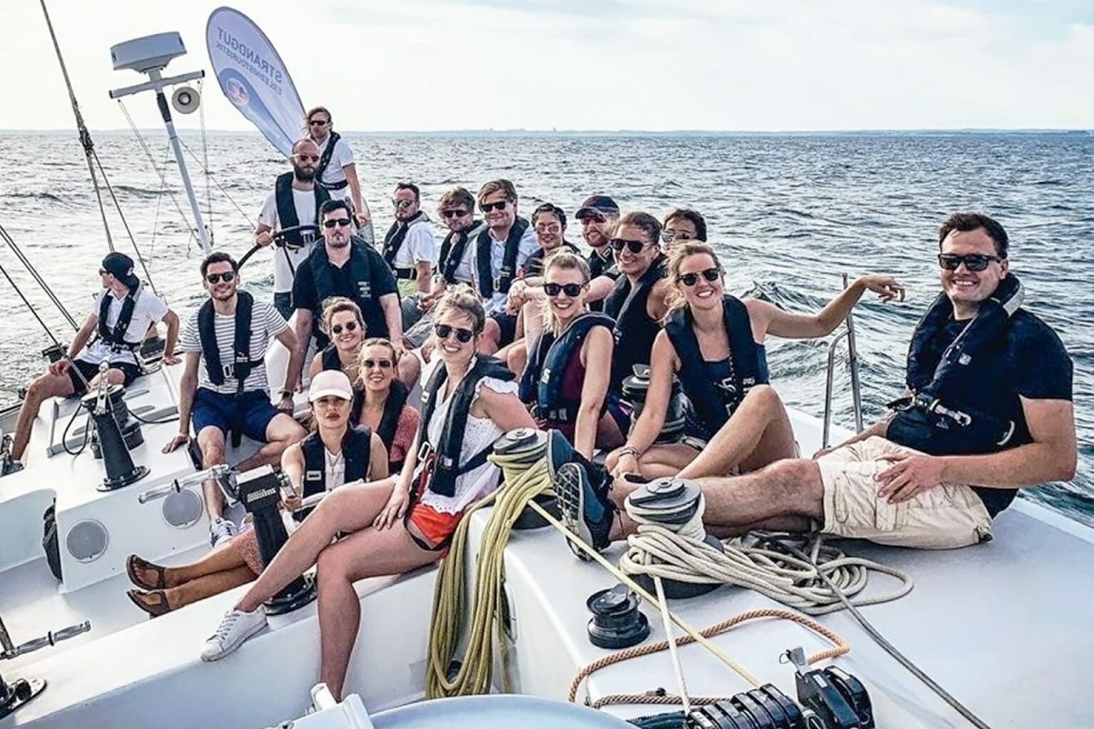 A large group of smiling people in casual clothing and life jackets sit on a sailboat deck on a sunny day, with the ocean in the background, reflecting the vibrant and collaborative spirit found at Spryker careers.