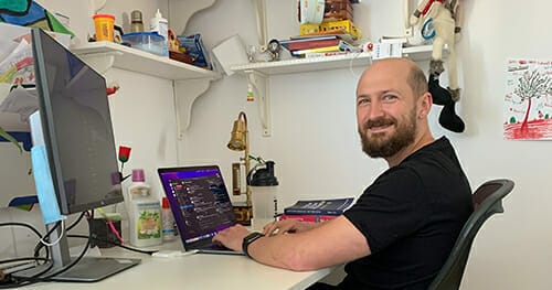 A man with a beard sits at a desk working on a laptop, exploring Spryker careers. An external monitor, books, and various office supplies are on the desk and shelves behind him.