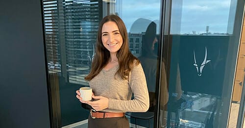 A woman with long brown hair, wearing a beige sweater and holding a white mug, stands in front of glass windows with city buildings in the background, considering Spryker careers for her next professional move.