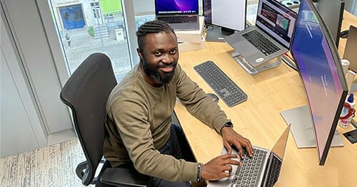 A person wearing a green sweater types on a laptop at a desk with multiple screens in an office setting, perhaps exploring opportunities on Spryker careers.