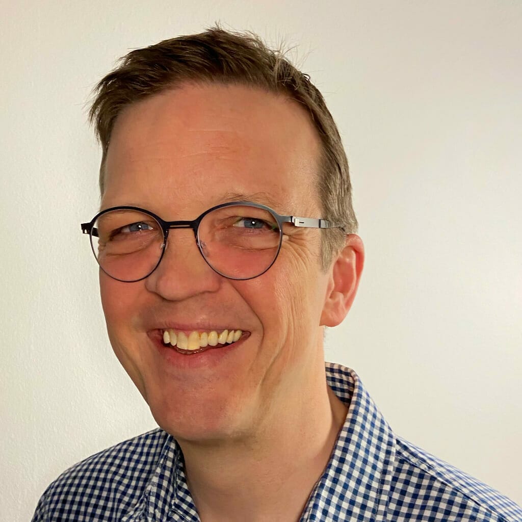 A person with short hair, smiling, wearing round glasses and a checkered shirt, stands against a plain white background, ready for Seamless Dubai 2022.