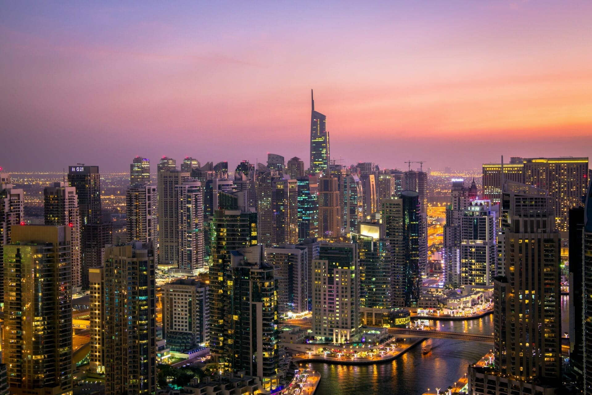 Aerial view of a cityscape at dusk, featuring numerous illuminated high-rise buildings and a winding waterway. The sky is a gradient of warm colors, reminiscent of Seamless Dubai 2022's vibrant energy.