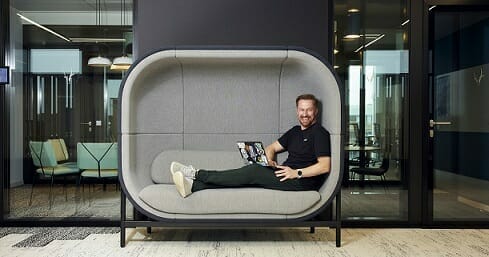 A man smiles while sitting with a laptop on a modern, cushioned sofa in an office environment with glass walls, embodying the dynamic and innovative spirit of Spryker careers.