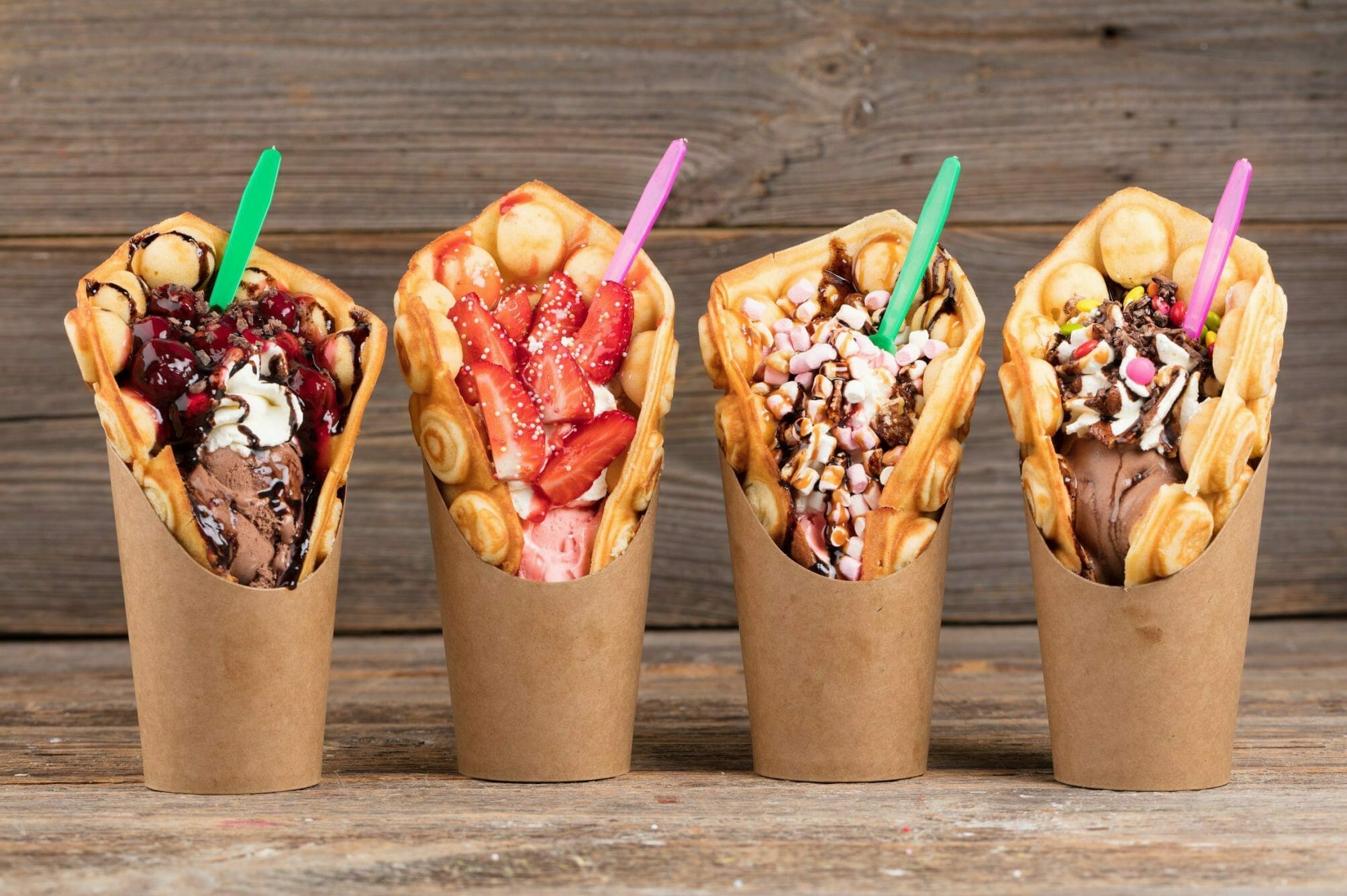 Four delightful waffle cone desserts, topped with an array of treats like chocolate, strawberries, and marshmallows, served in brown paper cups on a wooden surface. A sweet reminder of the fun and flavorsome moments from K5 Konferenz 2022.