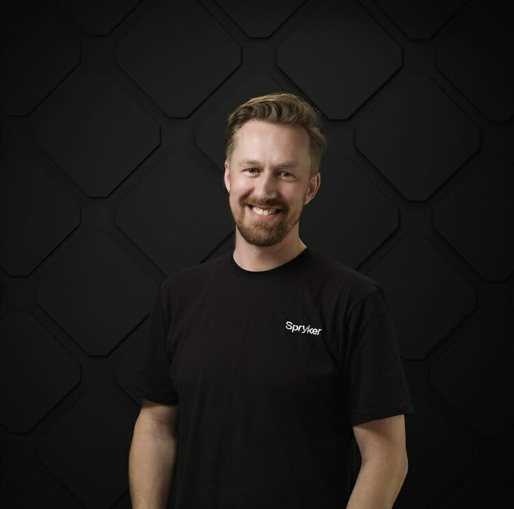 A man with a beard and mustache, wearing a black Spyker t-shirt, smiles in front of a dark, geometric background, looking like he's just finished giving one of his exciting press releases.