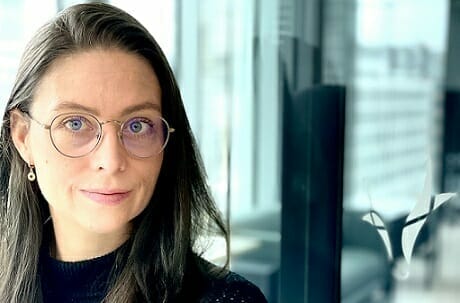 A woman with long dark hair and glasses stands near a window, wearing a black top and a faint smile, exuding the confident allure of someone who thrives in Spryker careers.