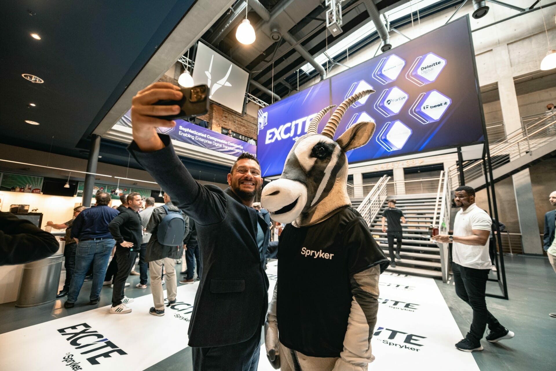 A man takes a selfie with someone in a goat costume at an event, standing on a floor with "Excite" written on it and a large sign displaying the same in the backdrop. The scene is filled with energy from the 2022 Spryker EXCITE takeaways.