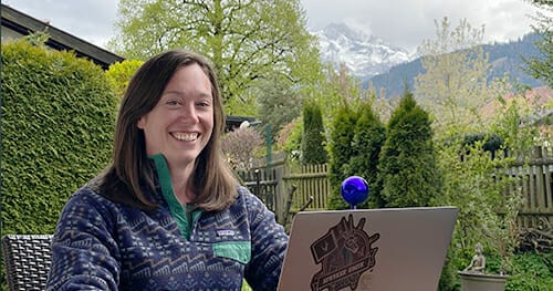 Person smiling, sitting outside with a laptop displaying stickers. Green trees and mountainous landscape are visible in the background. Discover opportunities with Spryker careers and work from inspiring locations like this.