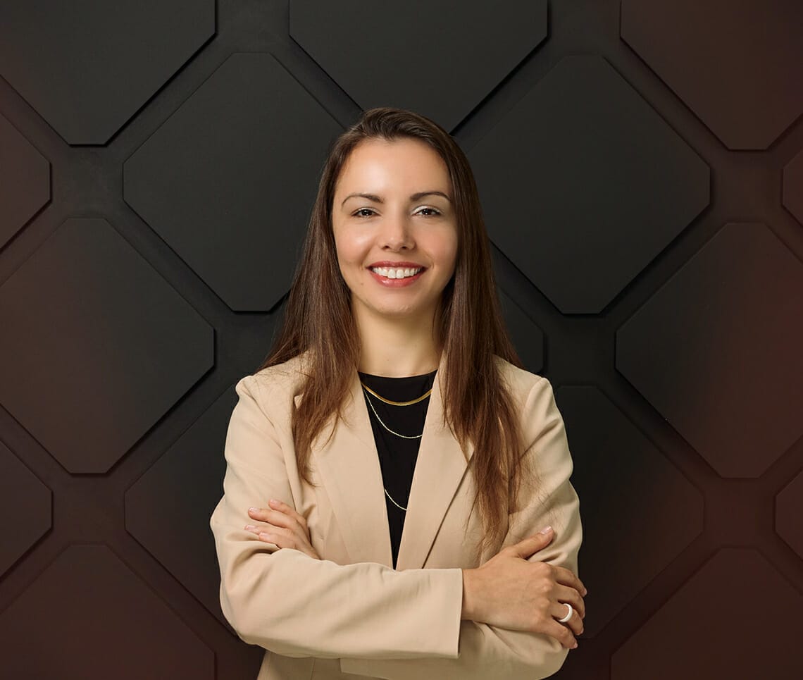 Woman smiling with arms crossed, wearing a beige blazer over a black top, standing in front of a geometric, dark-paneled background, ready to discuss the Customer Roadmap during the March 2023 webinar.