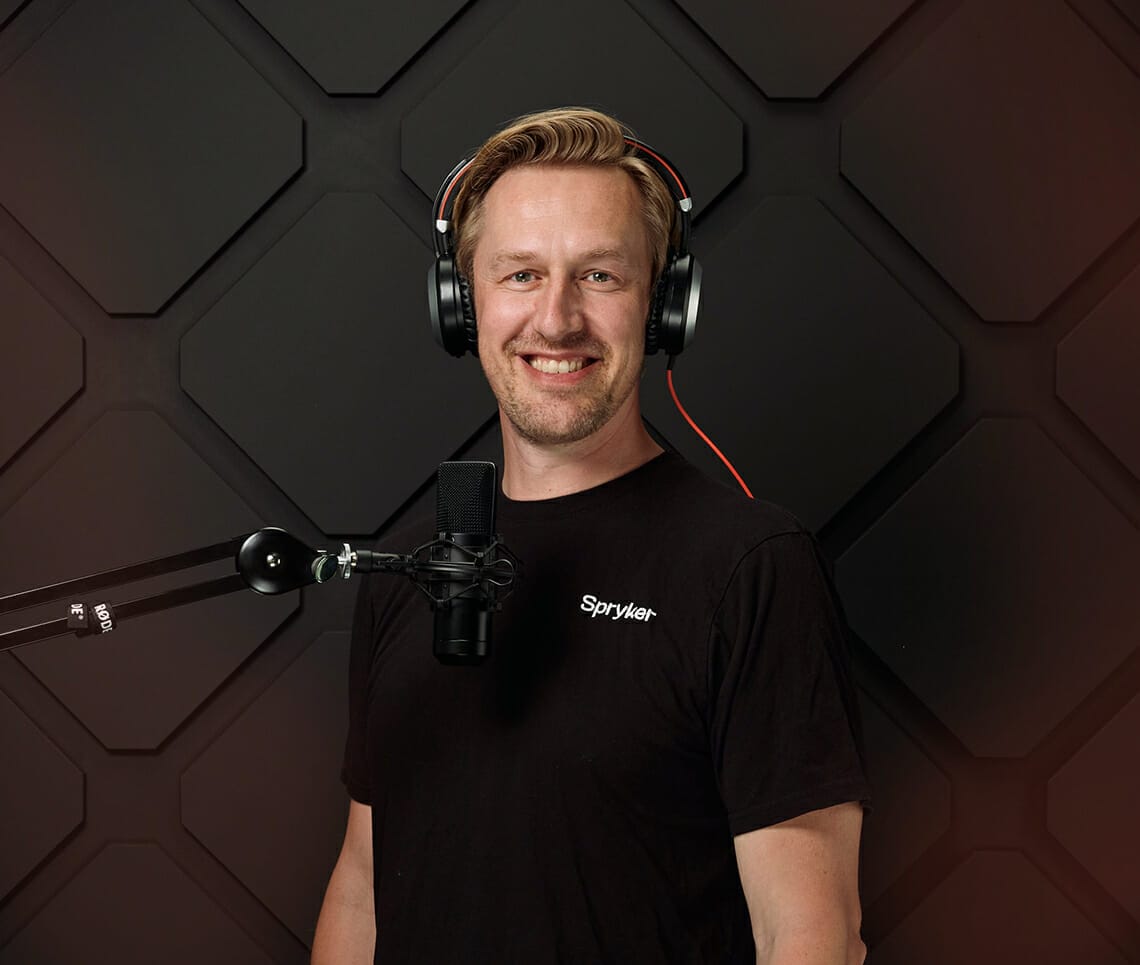 A person wearing a black t-shirt and headphones stands in front of a microphone with a geometric-patterned background, ready to go onair.