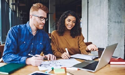 Two people, one in a blue shirt and the other in a brown shirt, are working together at a desk with a laptop, notepads, papers, and discussing strategies for b2b marketplaces.