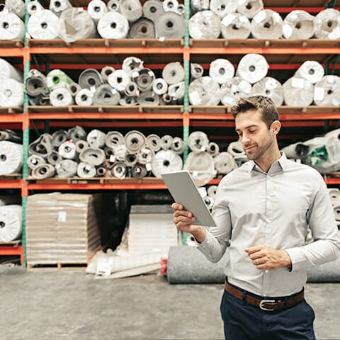 A man stands in a warehouse filled with large rolls of fabric or materials on shelves. He is looking at a tablet and appears to be checking inventory or data. He is wearing a light gray shirt and dark pants.