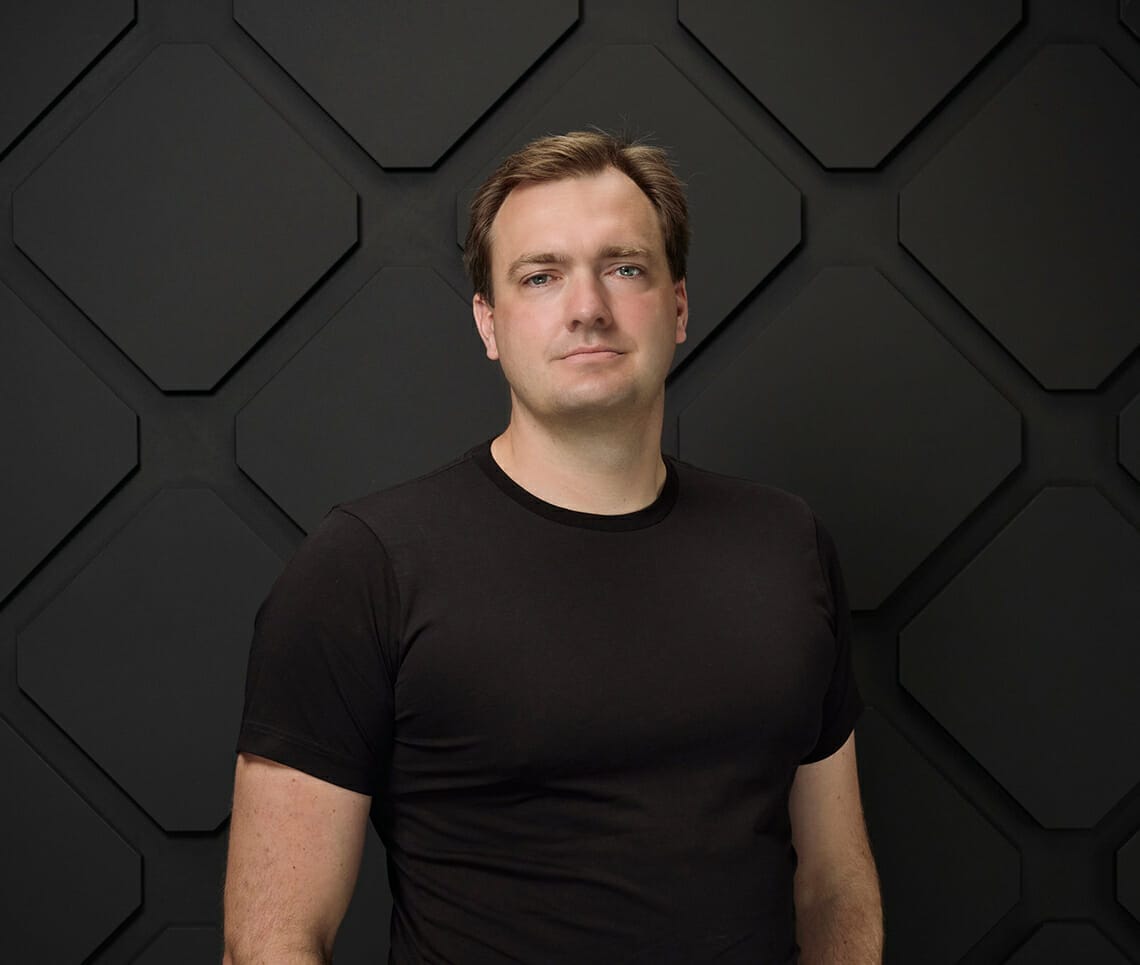 A man in a black t-shirt stands in front of a geometric-patterned, dark wall, looking at the camera, as if he's ready to go onair.