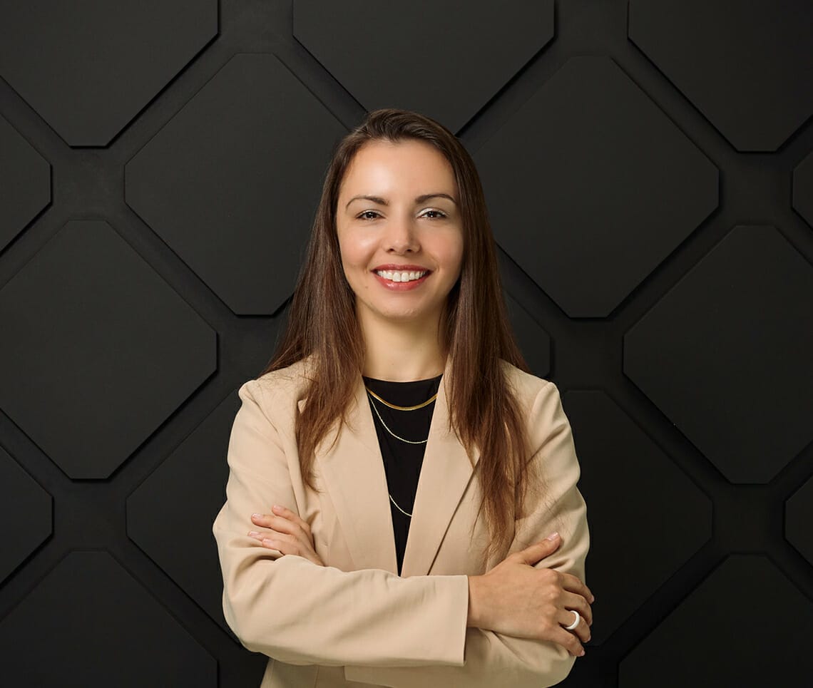 A woman with long brown hair stands confidently, arms crossed, wearing a light beige blazer in front of a black wall with a geometric pattern. Her poised demeanor embodies the essence of being onair.