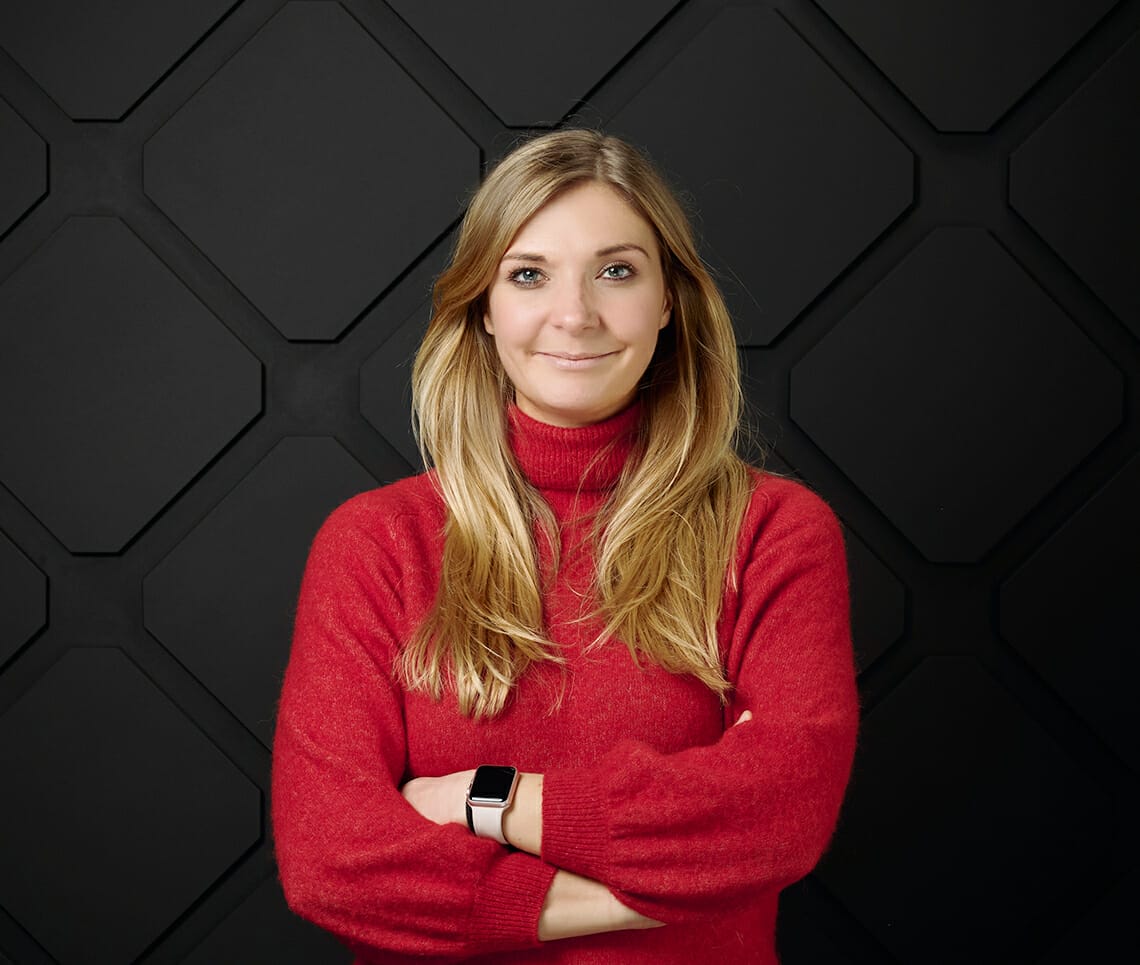 A woman with long blonde hair, wearing a red turtleneck sweater, stands with her arms crossed in front of a textured black background, looking onair ready.