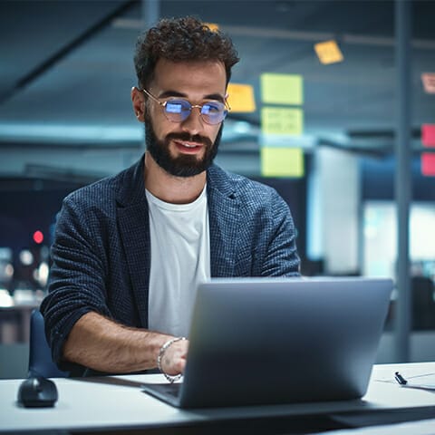 A man with glasses and a beard is sitting at a desk in an office, working on a laptop with composable commerce tools. There are sticky notes on the glass wall behind him.