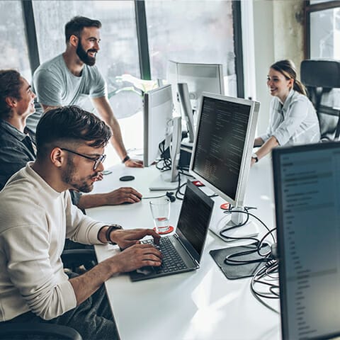 A group of people work and converse at desks with computers in an office setting. One person types on a laptop while others are engaged in a discussion about composable commerce solutions.