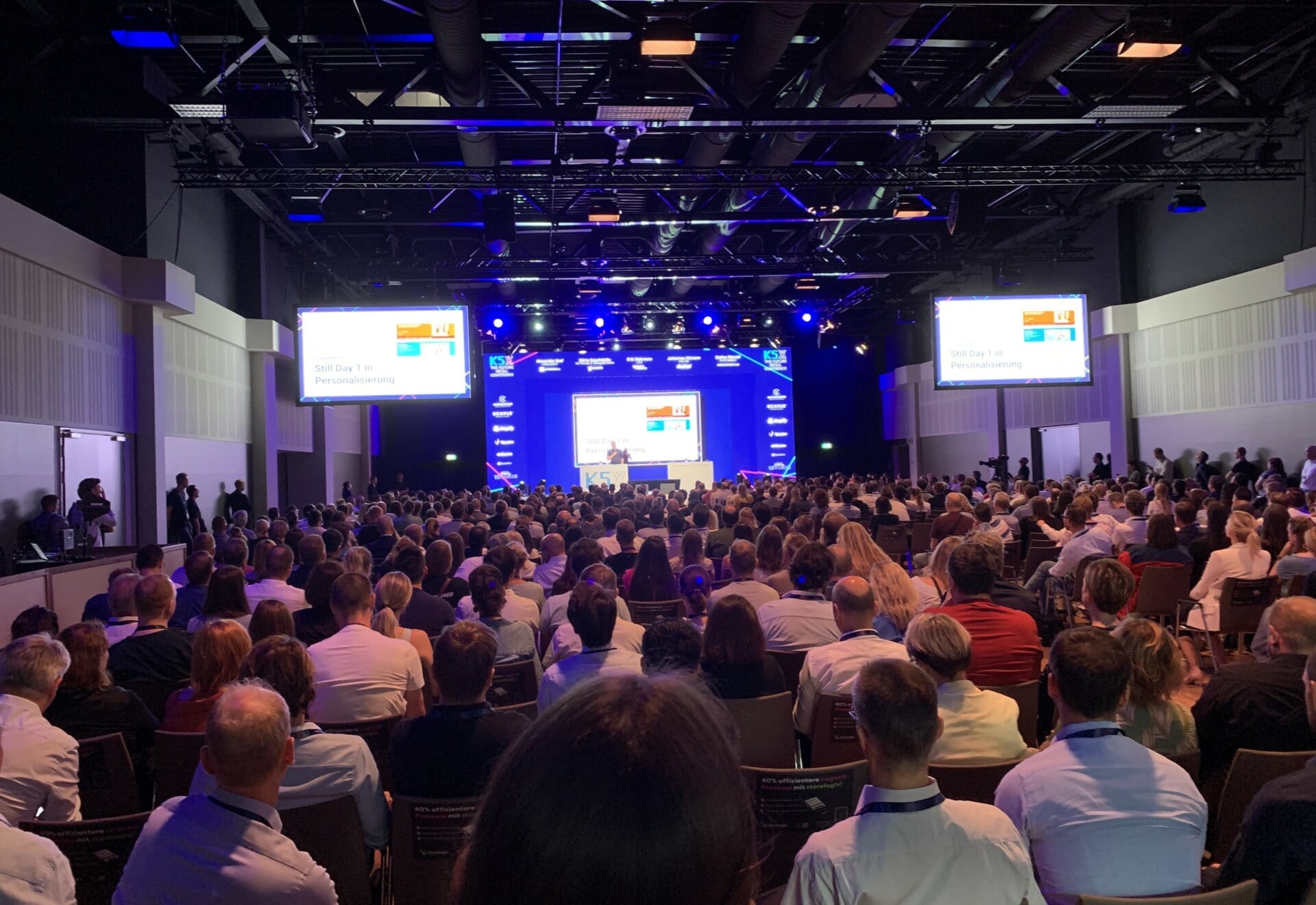 A large crowd watches a speaker on stage at a K5 Lehren conference. Two screens display slides on either side of the stage in a dimly lit auditorium.