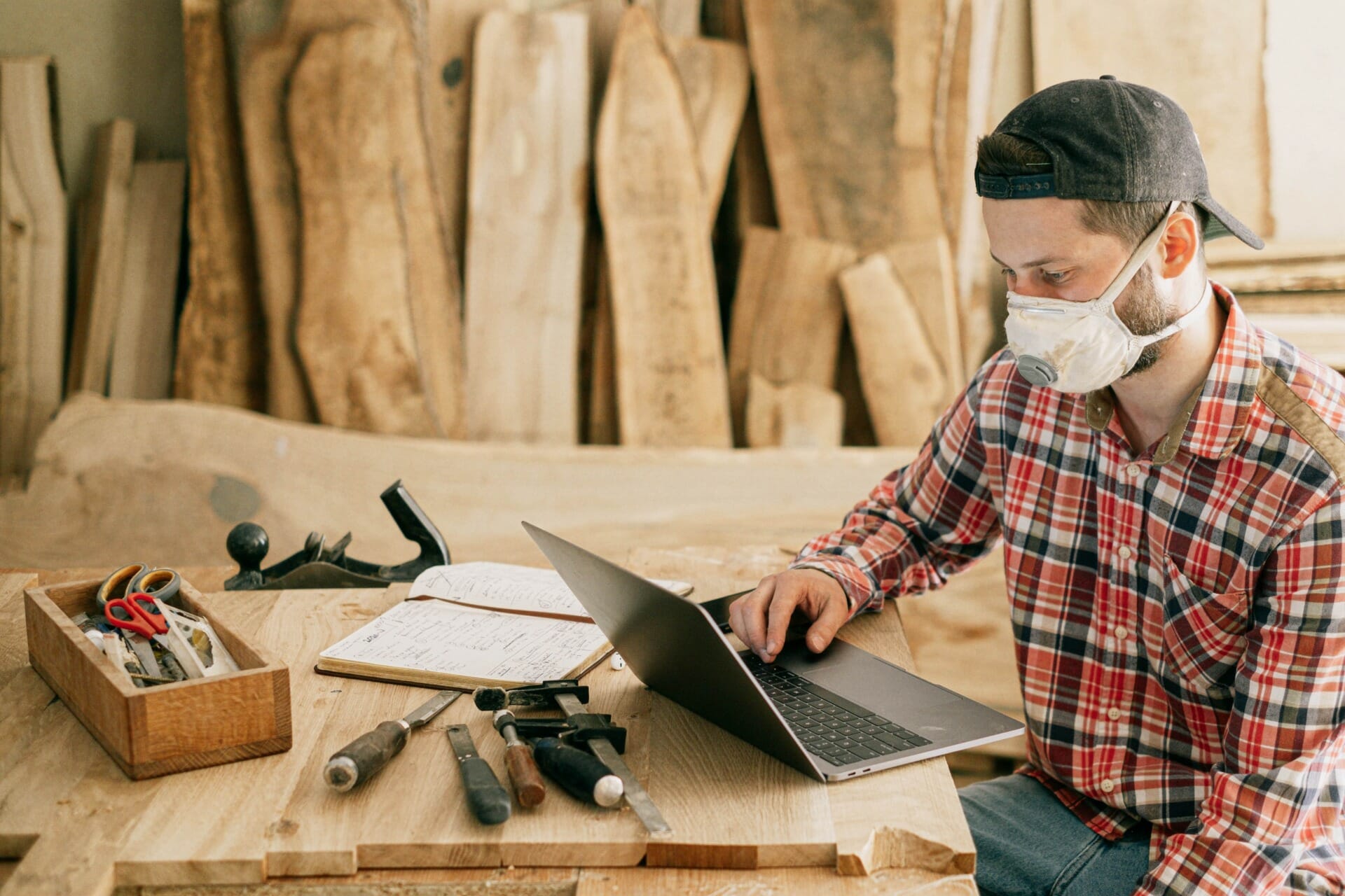 A person in a plaid shirt and a mask uses a laptop in a woodworking shop, with tools and wooden planks visible in the background, possibly researching D2C benefits for manufacturers.