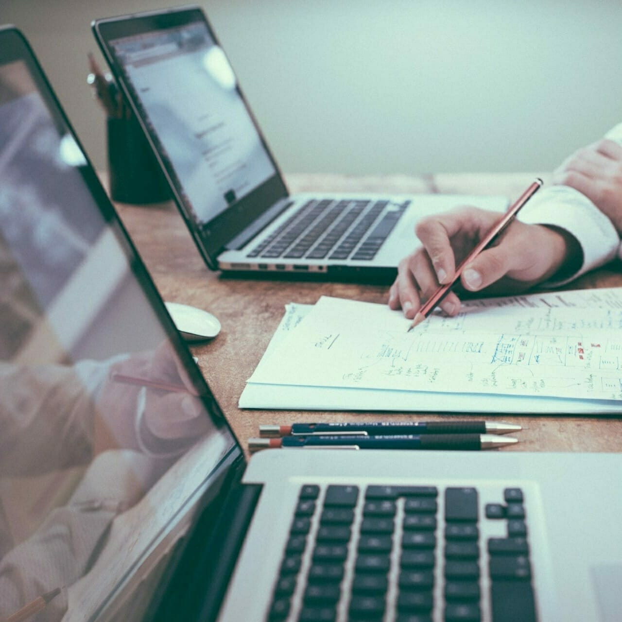 Two laptops on a wooden table, with a person's hand holding a pencil over a sheet of paper filled with handwritten notes and diagrams, possibly drafting the ultimate market guide.