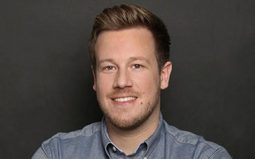 Smiling man with short brown hair and a beard, wearing a blue button-down shirt against a plain dark background, ready to discuss B2B solutions with Bloomreach and Spryker.