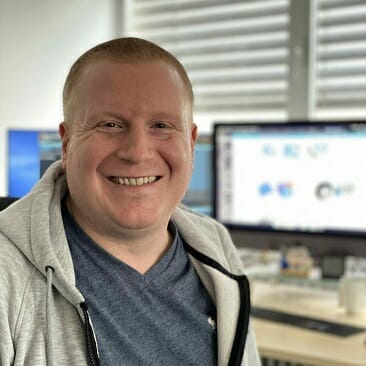 A person with short hair and a grey hoodie is smiling in front of a computer monitor, which is displaying various Oracle Commerce applications.