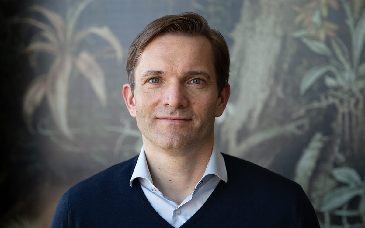 A person with short brown hair, wearing a white shirt and dark-colored sweater, stands in front of a leafy background, possibly attending the B2B-Wiesn event.