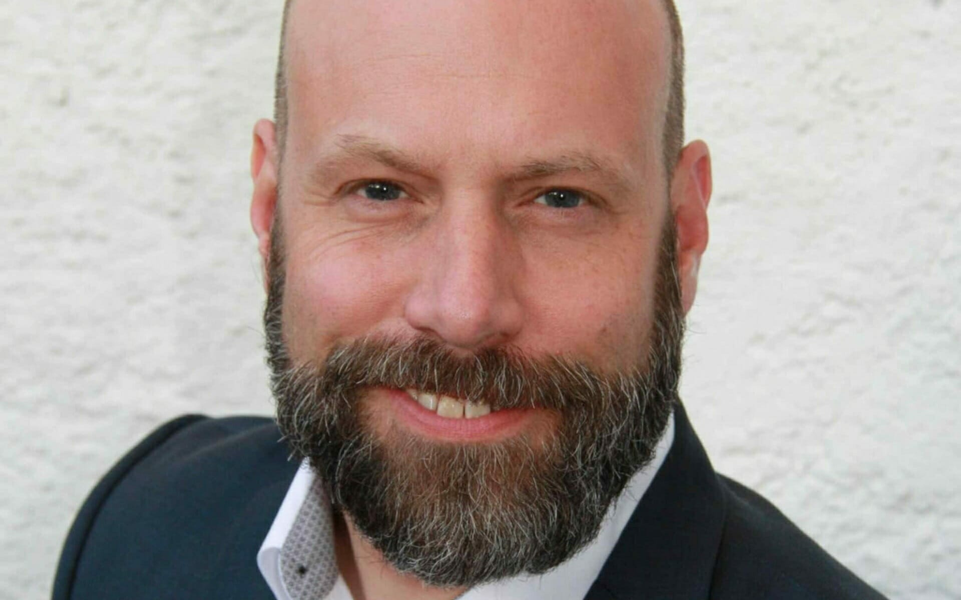 A man with a beard and bald head is smiling against a white textured background, embodying the spirit of b2b-wiesn. He is wearing a dark blazer over a white shirt.