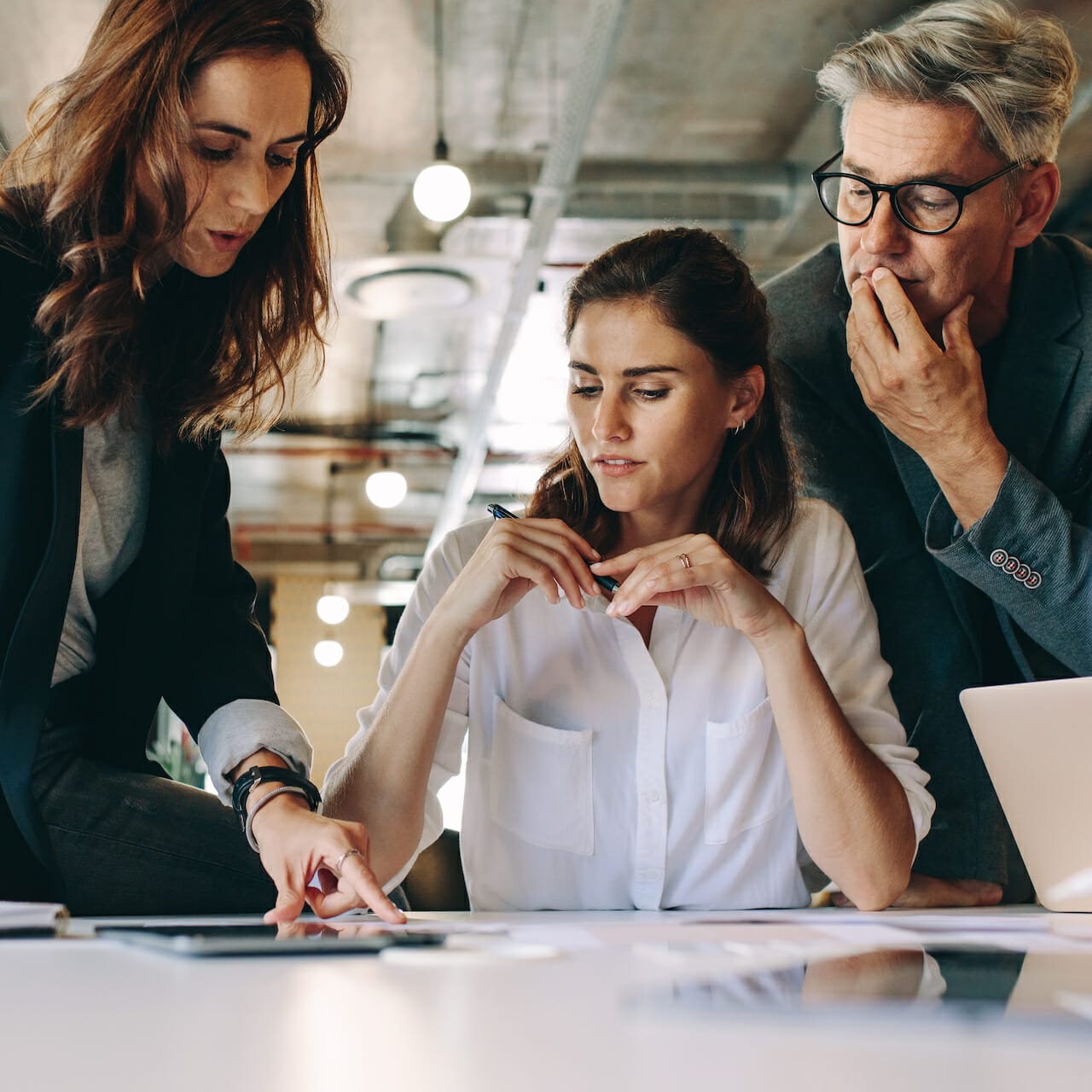 Three colleagues discuss work in a modern office. A woman on the left points at a document, the woman in the middle looks at it pensively, and the man on the right observes with a hand on his chin, likely considering the implications of channel conflict in their D2C strategy.