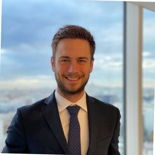 A man in a suit and tie stands smiling in front of a window with an urban skyline and cloudy sky in the background, looking poised for his presentation at e-show Madrid.